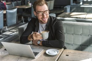 Close up of smiling freelancer sitting in cafe, drinking fresh coffee and using laptop