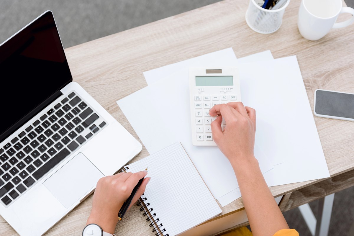 person using a calculator, notepad and laptop to manage finances